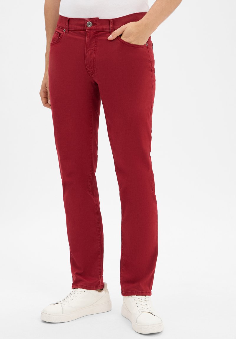 Jean en mélange de coton rouge avec une coupe ajustée, cinq poches et une fermeture à bouton, associés à des baskets blanches.