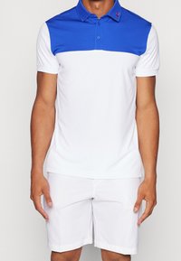 Pikétröja med blå övre del och vit nedre del, med krage och knapprad. Bärs med vita shorts. Slät tyg.