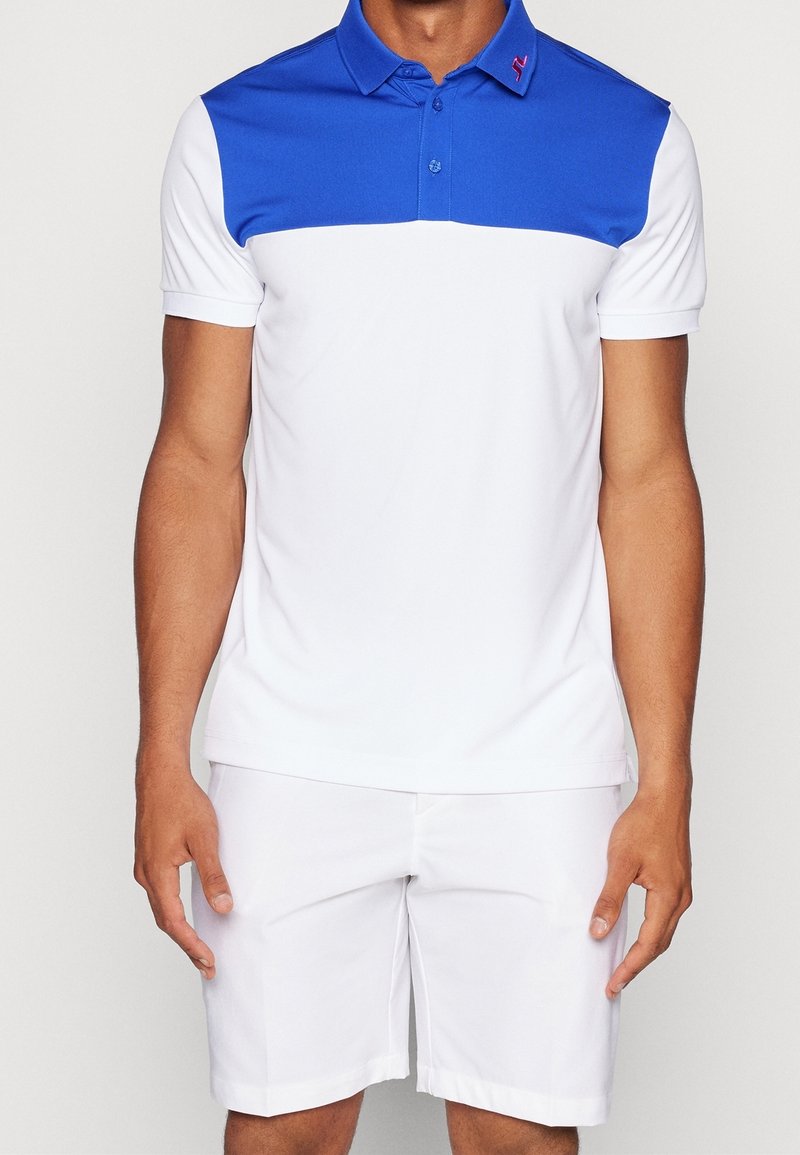 Pikétröja med blå övre del och vit nedre del, med krage och knapprad. Bärs med vita shorts. Slät tyg.