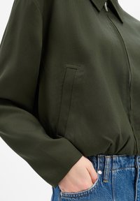 Olijfgroene bomberjack met ritssluiting, voorzien van een kraag en zijzak. Zachte, gestructureerde stof, gecombineerd met blauwe high-waisted denim.