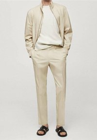 Beige letvægtsjakke med lynlås, cremefarvet ribbet sweater, beige bukser og sorte sandal med remme. Glat stof med skræddersyet pasform.