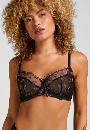 Frau mit lockigem Haar, die einen schwarzen transparenten BH mit lila Blumenstickerei und dünnen Trägern vor einem einfarbigen hellen Hintergrund trägt.
