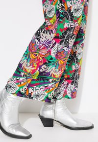 Kleurige wijde broek met een gedurfde, grafische print van bloemen en gezichten, gecombineerd met glanzende zilveren enkellaarzen met een lage hak.