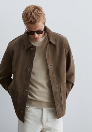 Joven con gafas de sol oscuras, chaqueta de ante marrón, suéter beige y pantalones blancos, mirando hacia abajo con las manos en los bolsillos de la chaqueta.