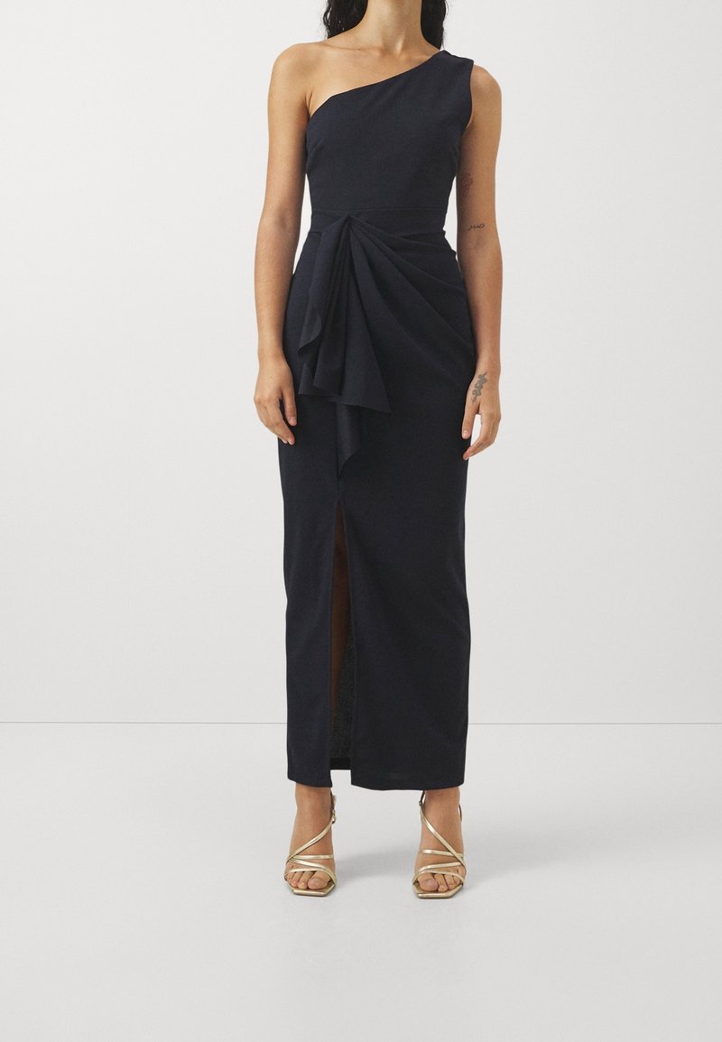 Femme portant une longue robe noire asymétrique avec un détail drapé sur le devant et une fente haute à la cuisse, associée à des talons hauts dorés à brides.