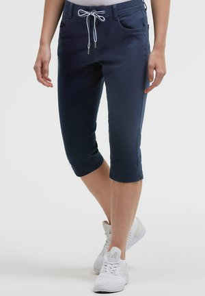 Person wearing navy blue cropped pants with drawstring waist and white sneakers, standing against a plain background.