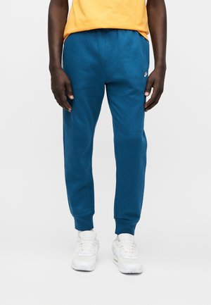 Osoba oblečená v modrých teplácích Nike, bílých teniskách a žlutém tričku, stojící proti jednoduchému světlému pozadí.