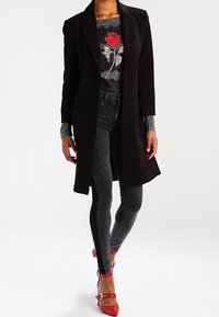 Abrigo largo negro sobre una camiseta gris con un diseño de rosa roja, combinado con leggings gris oscuro y zapatos de tacón rojo con correas en el tobillo.