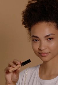 Creamy concealer in a beige tube with a black cap, held by a person with curly hair, wearing a white shirt with pearl accents.