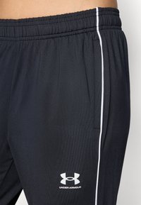 Čierne športové nohavice vyrobené z hladkej tkaniny, s bielym bočným pruhom a logom Under Armour blízko lemu. Elastický pás.