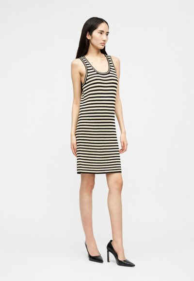 Ženska stoji, oblečena v črno-belo črtasto brezrokavno obleko do kolen in črne čevlje s peto, na enostavni podlagi.