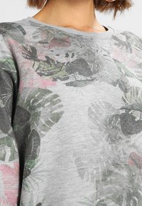 Zbliżenie na osobę w szarym swetrze z wyblakłym tropikalnym wzorem w odcieniach zieleni, czerni i różu.