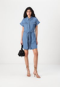 Denim obleka s kratkimi rokavi, gumbnim zapiranjem spreda, pasom, frčenim robom in dvema žepoma na prsih. Usklajena z črno platneno torbo.