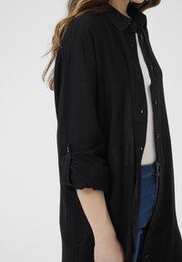 Czarna koszula z guzikami, z kołnierzykiem, długimi rękawami podwiniętymi na mankietach, wykonana z lekkiego materiału, w zestawie z białą bluzką i niebieskimi dżinsami.