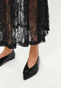 Sort lace nederdel med blomster mønstre, semi-gennemsigtige lag og en bølget kant. Sorte spidse flade sko synlige nederst.