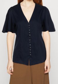 Blusa navy con scollo a V, maniche corte a palloncino e chiusura frontale con bottoni. Presenta un orlo dritto e una trama di tessuto liscia.