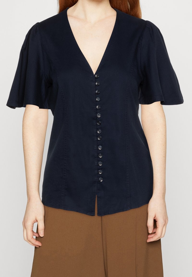 Blusa navy con scollo a V, maniche corte a palloncino e chiusura frontale con bottoni. Presenta un orlo dritto e una trama di tessuto liscia.
