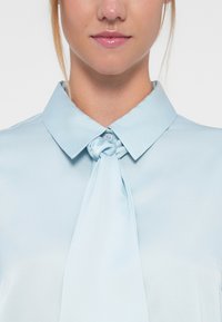 Blusa azul clara com colarinho e detalhe de laço atado no pescoço. Tecido suave e macio com um ligeiro brilho. Textura subtil e fecho de botão.
