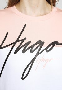 Close-up van een persoon die een roze en witte top draagt met grote zwarte en kleinere perzikkleurige 'Hugo' scriptlogo's over de borst.