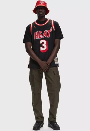 Uomo con una maglia da basket nera dei Miami Heat con il numero 3, pantaloni cargo verdi, scarpe da ginnastica nere e un cappello a secchiello rosso.