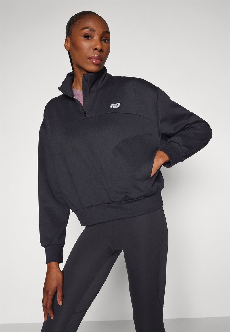 Donna che indossa un felpa nera New Balance con zip a quarti e leggings abbinati, posando con una mano in tasca su uno sfondo semplice.