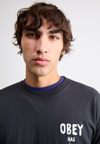 Sweatshirt negru din bumbac cu guler rotund, având textul alb "OBEY HAS" pe pieptul stâng. Sub acesta, se vede un guler albastru.