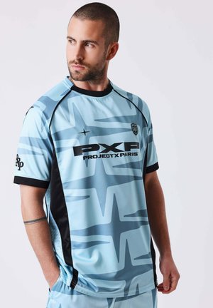 Uomo con capelli corti e barba che indossa una maglia sportiva azzurro chiaro Project X Paris con motivo astratto, che guarda verso sinistra.