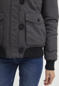 Grå pufferjakke med fem knapper, to frontlomme med klaff, og ribbestrikkede mansjetter. Strukturert stoff, hofte-lengde design, og en slim passform i midjen.