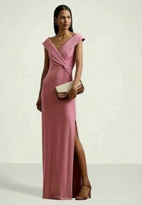 Ροζ, εφαρμοστό βραδινό φόρεμα με καπέλα μανίκια, λεπτομέρεια στριφτού κορσέ και πλάγιο σκίσιμο. Συνδυάζεται με μια μπεζ τσάντα χειρός με αξεσουάρ με δίπλα. 