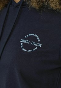 Felpa blu con cappuccio che presenta una grafica circolare con il testo: "NON È MAI TROPPO TARDI," "ATTUALMENTE - IN EVOLUZIONE," e "INIZIA ORA - GIOCA SOLO."