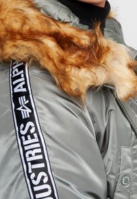 Vadderad grå jacka med en krage av konstpäls, med ett svart och vitt "ALPHA INDUSTRIES" logoband längs ärmen och en dragkedjeförsedd ficka.