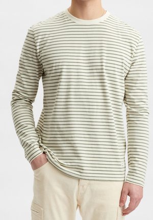Mand iført en cremefarvet langærmet skjorte med tynde grå horisontale striber og lyse beige bukser, den ene hånd i lommen, hvid baggrund.