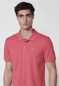 Roza polo majica z klasičnim ovratnikom, z dvema gumboma in majhnim logotipom na levem prsnem delu. Tkanina ima gladko teksturo.