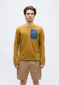 Νεαρός άνδρας που φοράει μουσταρδί κίτρινη ενδυμασία με υφή και μακρυμάνικη μπλούζα με μπλε τσέπη με φερμουάρ και μπεζ cargo σορτς, στέκεται μπροστά σε ένα απλό λευκό φόντο.