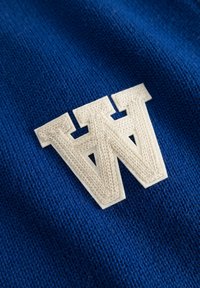 Tissu en maille bleue avec une lettre "W" blanche brodée et des détails texturés. Le tissu est doux et finement tissé.