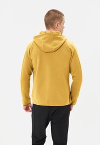 Felpa gialla in pile con cappuccio e dettagli neri, caratterizzata da una texture liscia. La persona indossa pantaloni neri ed è mostrata di spalle.