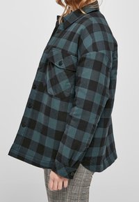 Camisa de cuadros en tonos verde azulado y negro, hecha de una tela suave. Cuenta con un collar, un bolsillo delantero y un corte relajado.