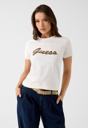 Jeune femme aux cheveux bruns lâchés porte un t-shirt blanc Guess et un jean bleu foncé, debout les mains dans les poches devant un fond uni.