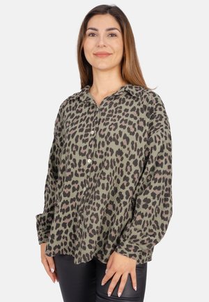 Femme portant une chemise ample à manches longues boutonnée, imprimé léopard vert et noir, associée à un pantalon noir, se tenant devant un fond blanc.