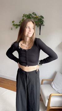 Črn rebrast dolg rokav krojen top s sprednjimi gumbi; v kombinaciji z visokim črnim širokim hlačami; ozadje vključuje sobno rastlino in pohištvo.