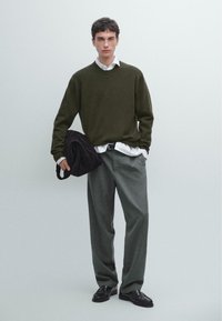 Maglione verde oliva sovrapposto a una camicia bianca, abbinato a pantaloni grigi chiari. Tiene una borsa marrone scuro e indossa scarpe nere.