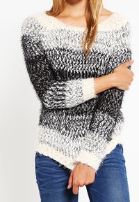 Eine Frau trägt einen flauschigen schwarz-weiß-grau gestreiften Pullover mit blauen Jeans, hat die Arme verschränkt und hält einen Arm.