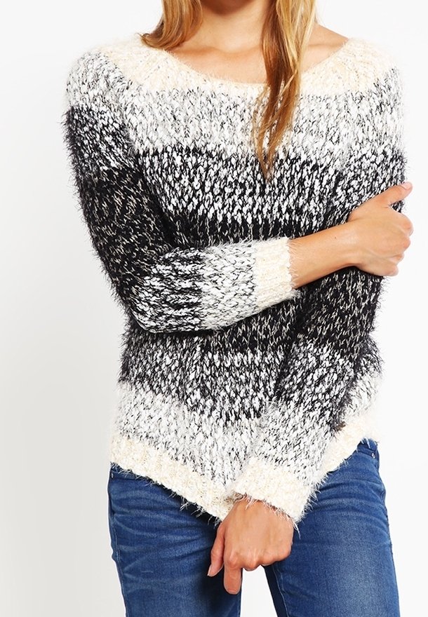 Eine Frau trägt einen flauschigen schwarz-weiß-grau gestreiften Pullover mit blauen Jeans, hat die Arme verschränkt und hält einen Arm.
