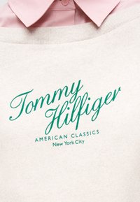 Μπεζ φούτερ με πράσινη γραφή "Tommy Hilfiger", τα λόγια "American Classics" και "New York City" πάνω από ένα ροζ πουκάμισο με γιακά.