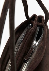 Sac en daim marron avec deux compartiments zippés, doté de quincaillerie en argent et d'une texture douce. Les poignées sont intégrées dans le design.