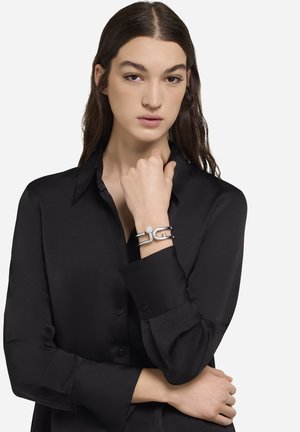 Bracelet en argent avec un logo proéminent, présentant un détail rond texturé, porté sur un poignet avec un chemisier noir. Finition lisse, design ouvert.