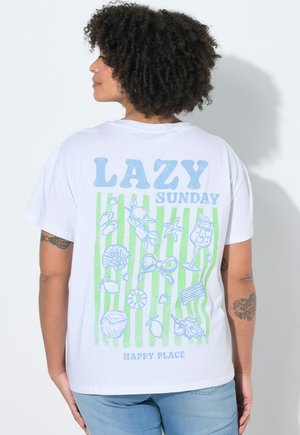Person mit lockigem Haar, seitlich blickend, trägt ein weißes T-Shirt mit grünen Streifen und blauen, strandbezogenen Illustrationen sowie dem Text "LAZY SUNDAY HAPPY PLACE."