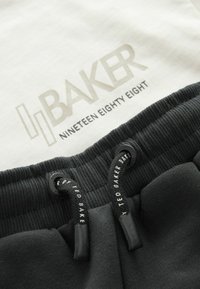 Gros plan sur une ceinture noire côtelée avec des cordons de serrage marqués Ted Baker et un tissu blanc affichant le texte "Ted Baker Nineteen Eighty Eight".