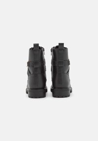 Paire de bottines noires avec des brides texturées et des fermetures éclair latérales, vues de dos sur un fond blanc uni.