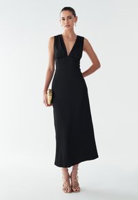 Melnas, bezroku maksi kleita ar dziļu V-veida izgriezumu, teksturētu audumu, piegulošu vidukli un plūstošu siluetu; kopā ar zeltainām sandales ar siksniņām.